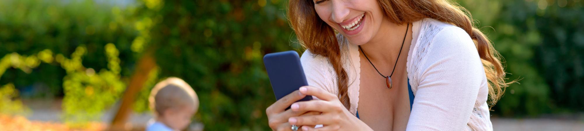 Mother in park smiling at cell phone with baby sitting on ground in background